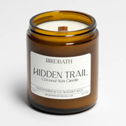A Birdbath Hidden Trail coconut wax candle in an amber glass jar with a white label, set against a clean white background.