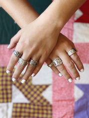 A close-up of two crossed hands wearing five silver-toned rings with ornate engraved patterns, set against a colorful quilted fabric background.