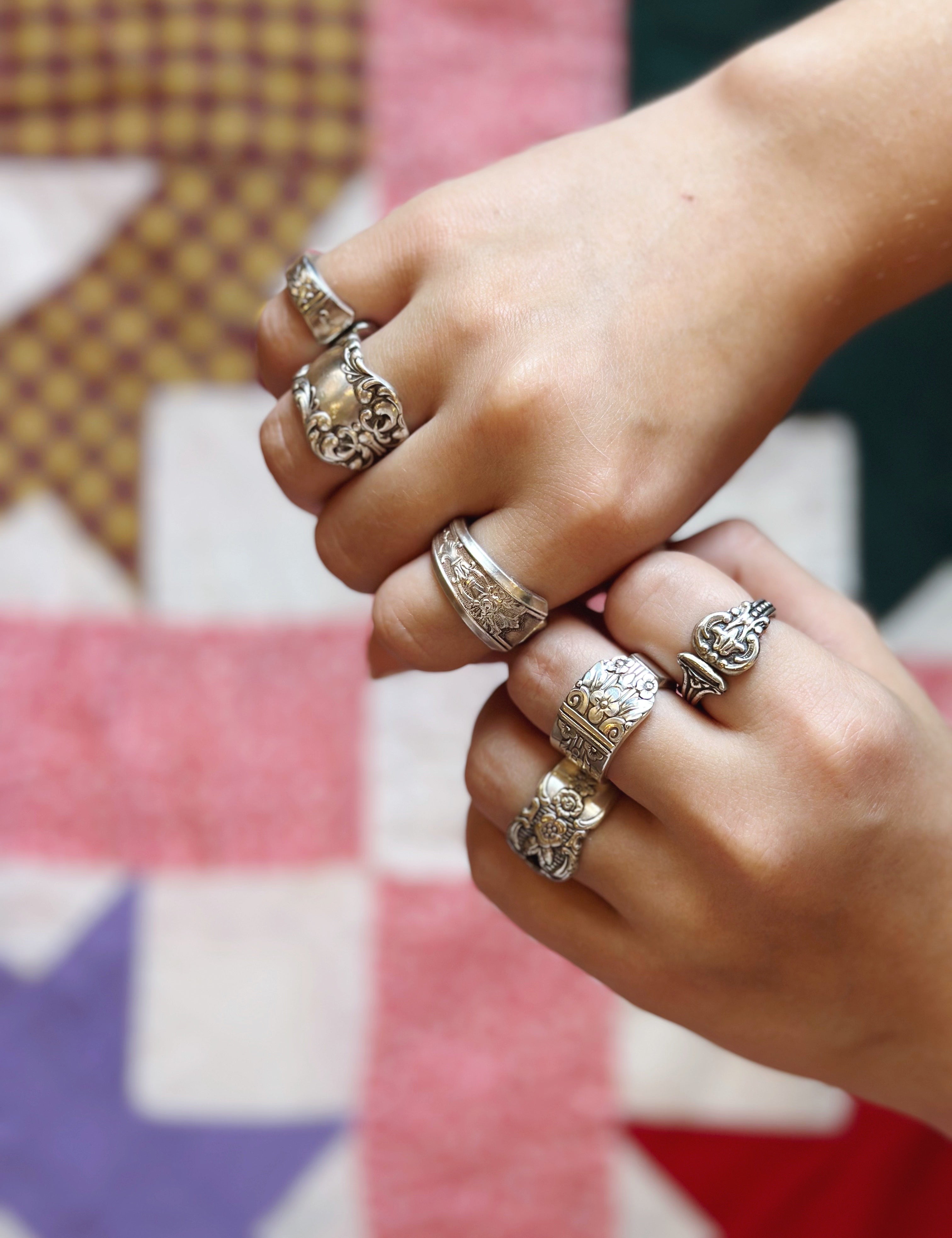 A close-up of two hands wearing multiple ornate silver rings with floral and scrollwork designs, set against a blurred multicolored quilt background.