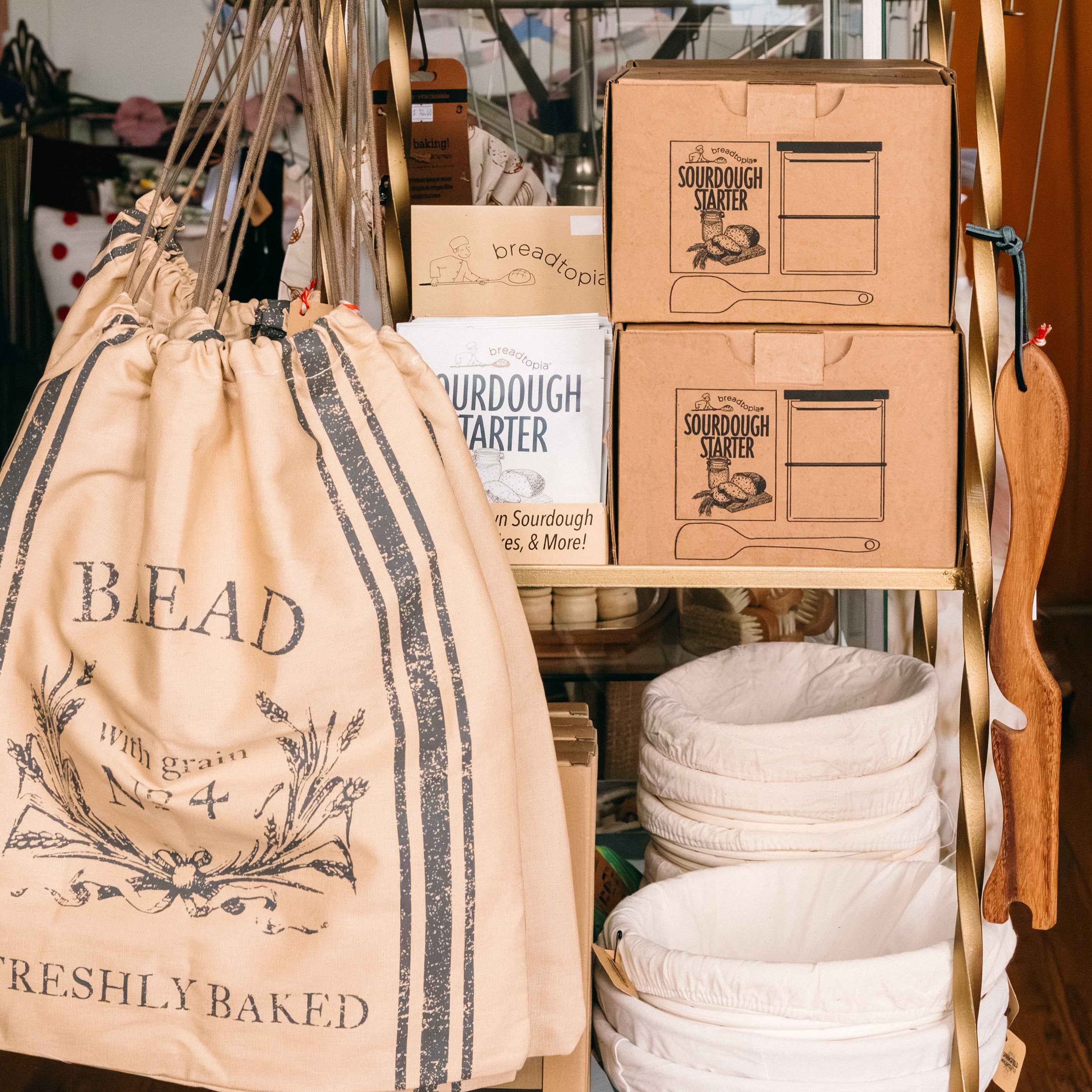 Bread-themed bags, sourdough starter kits, and lined baskets on a metal shelf.

Description: Categories image promoting Sourdough goods on the Springfield Mercantile Co. homepage. 
Keywords: Springfield Mercantile Co., sourdough, categories section, online shop, Shopify
